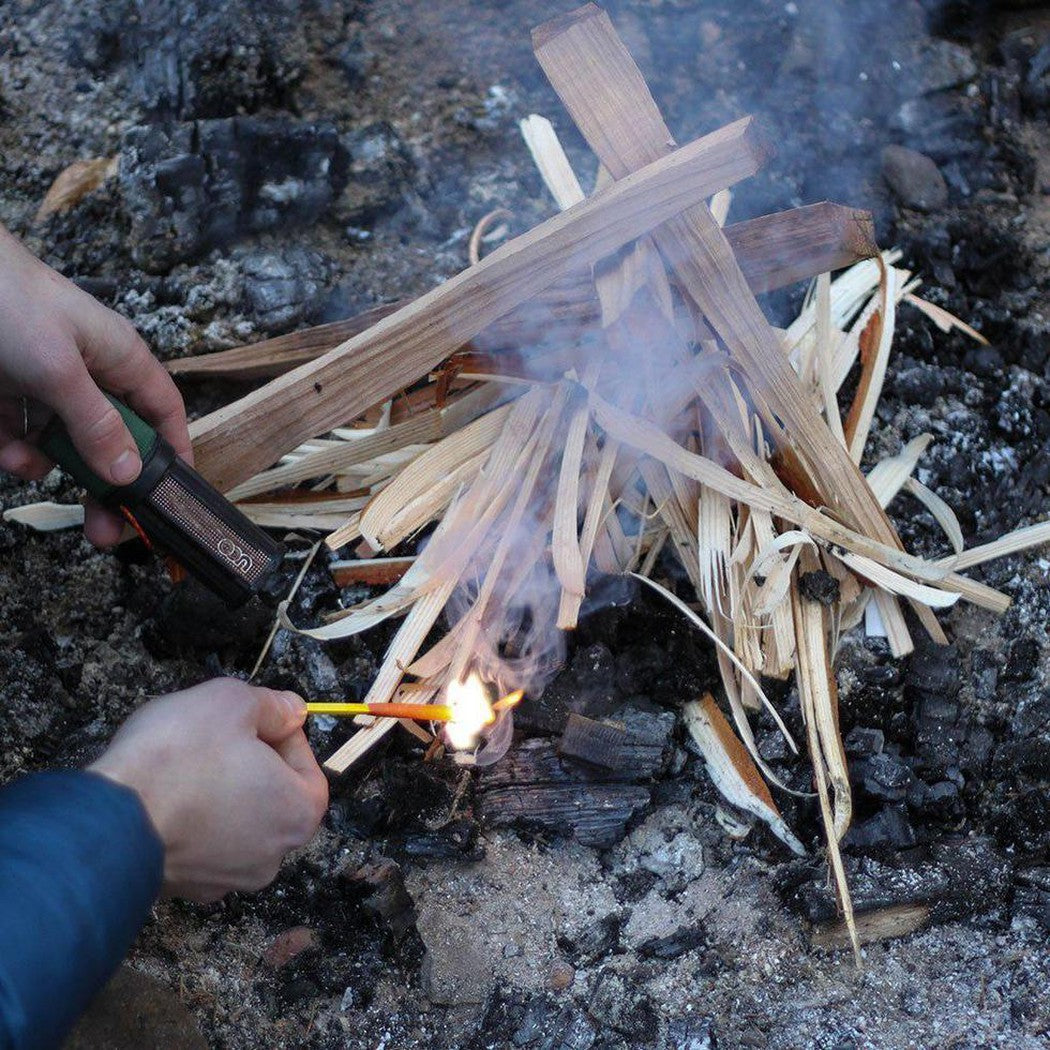 A person uses the UCO Titan Stormproof Match Kit to light a small fire with kindling in an outdoor setting.