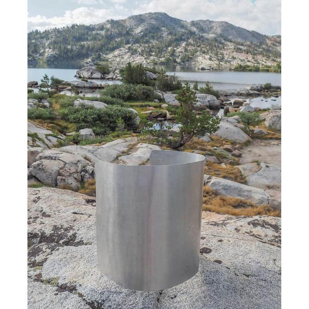 A stainless steel Ursack Aluminum Liner sits on a rocky surface with a lake and mountains in the background.
