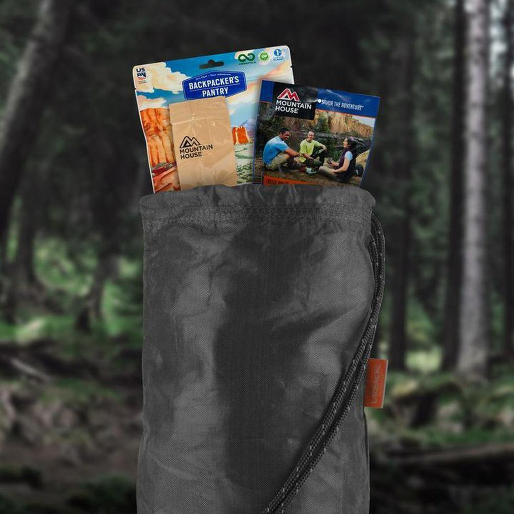 A charcoal Ursack Major food storage bag holds Backpacker's Pantry and Mountain House food packets against a forest background.