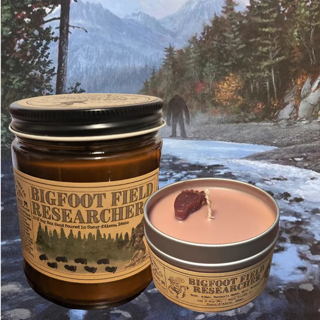 A jar and tin of Wanderlust Folk Bigfoot Field Researcher 4 oz candles sit on a forest path background with a bear silhouette.