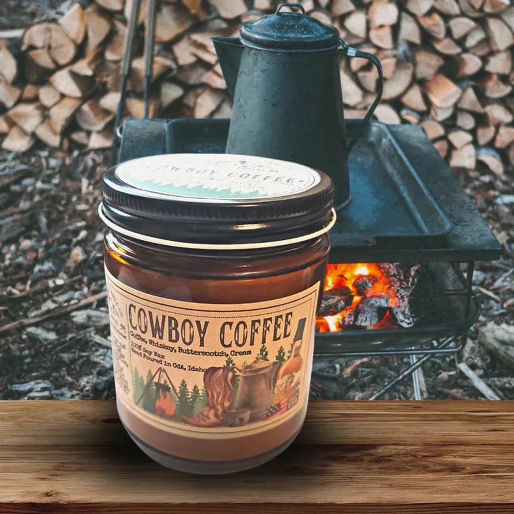 A 4 oz Wanderlust Folk Cowboy Coffee candle sits on a wooden surface with a campfire and stacked wood in the background.