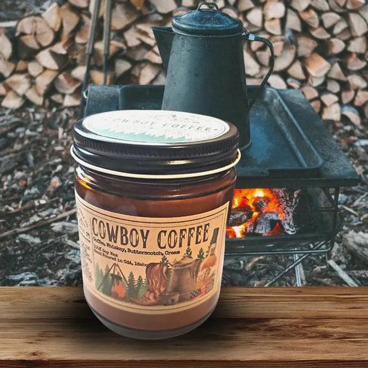 A 4 oz Wanderlust Folk Cowboy Coffee candle sits on a wooden surface with a campfire and stacked wood in the background.