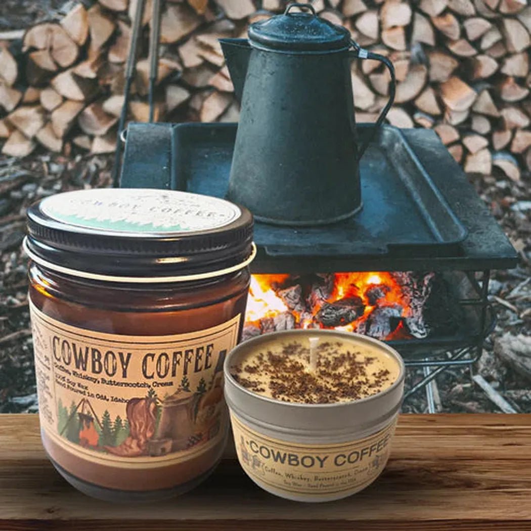 A 4 oz Wanderlust Folk Cowboy Coffee candle and tin sit on a wooden surface with a campfire and stacked wood in the background.