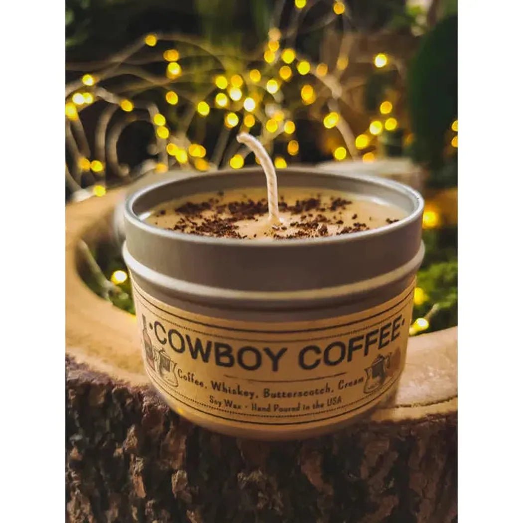 A 4 oz Wanderlust Folk Cowboy Coffee candle sits on a tree stump with twinkling lights in the background.