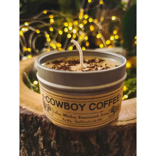 A 4 oz Wanderlust Folk Cowboy Coffee candle sits on a tree stump with twinkling lights in the background.