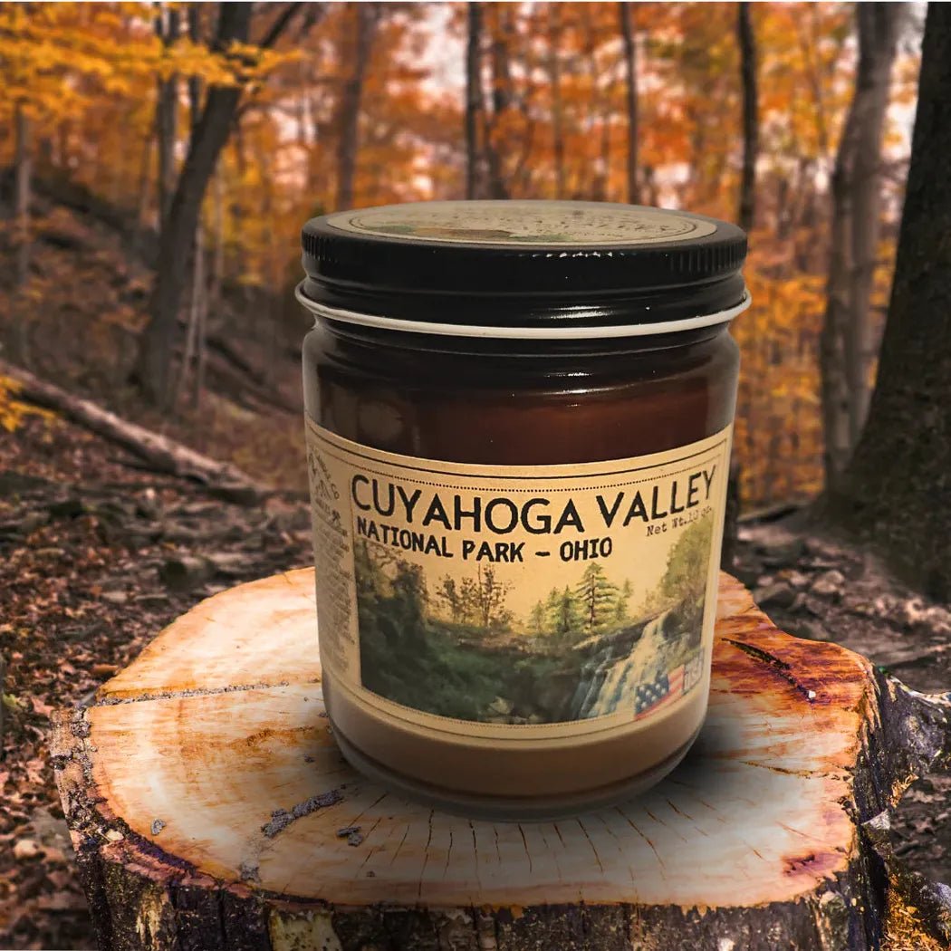 A 4 oz Wanderlust Folk Cuyahoga Valley candle sits on a tree stump in a forest with autumn leaves.