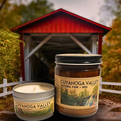Two Cuyahoga Valley candles sit in front of a red covered bridge with trees.