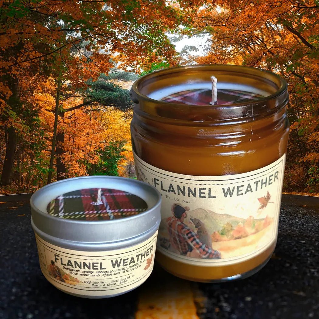 Two Flannel Weather candles sit on a dark surface with an autumn forest background.