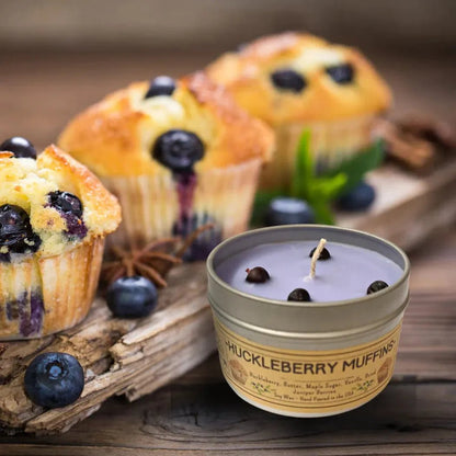A close-up of a Wanderlust Folk Huckleberry Muffins 4 oz candle tin next to blueberry muffins on a wooden board.