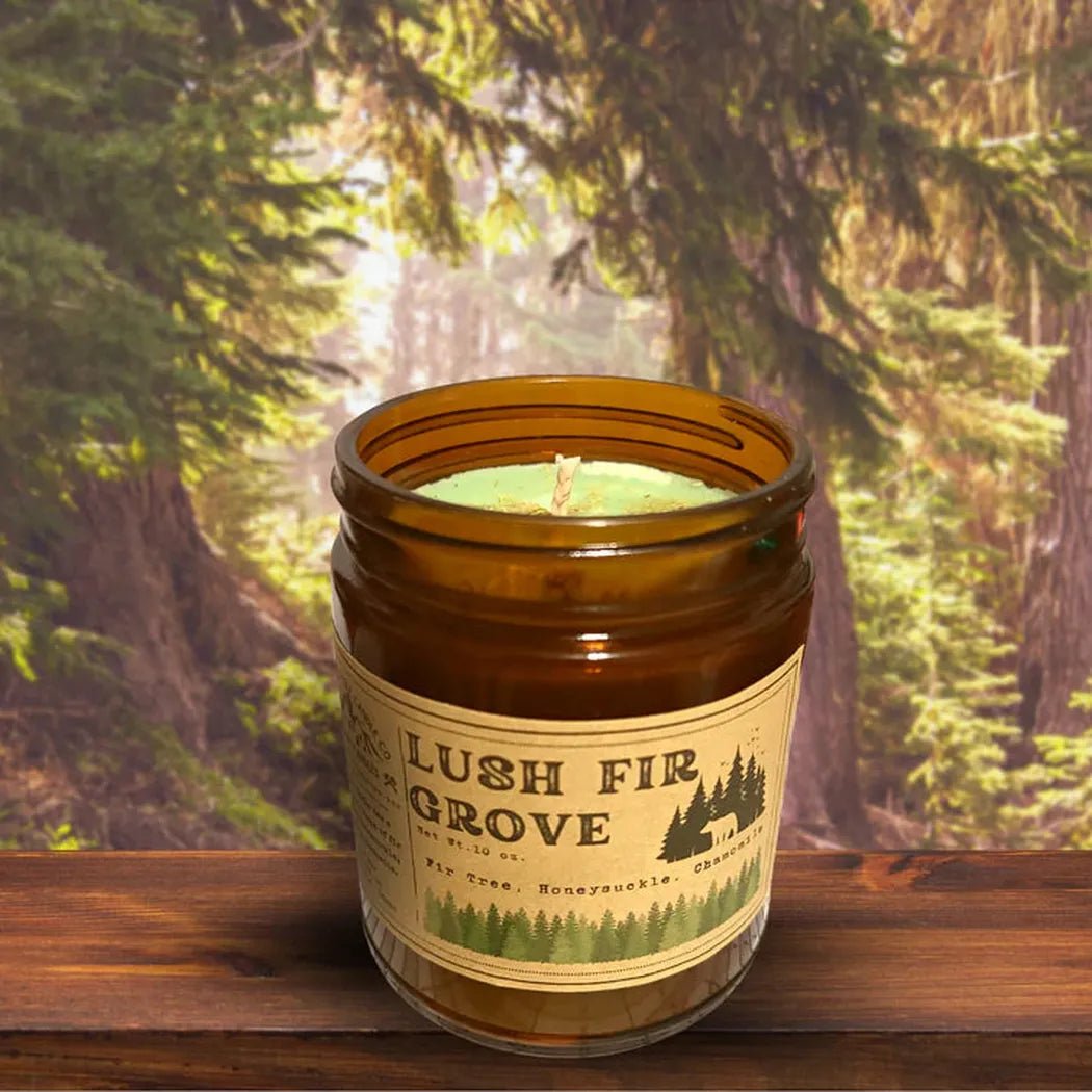 A 4 oz Wanderlust Folk Lush Fir Grove candle sits on a wooden surface with a forest background.