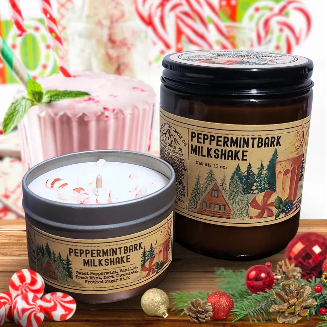 Two peppermint bark milkshake candles, one 4 oz in a charcoal tin and one larger jar, sit on a wooden surface with Christmas decorations and a peppermint milkshake in the background.