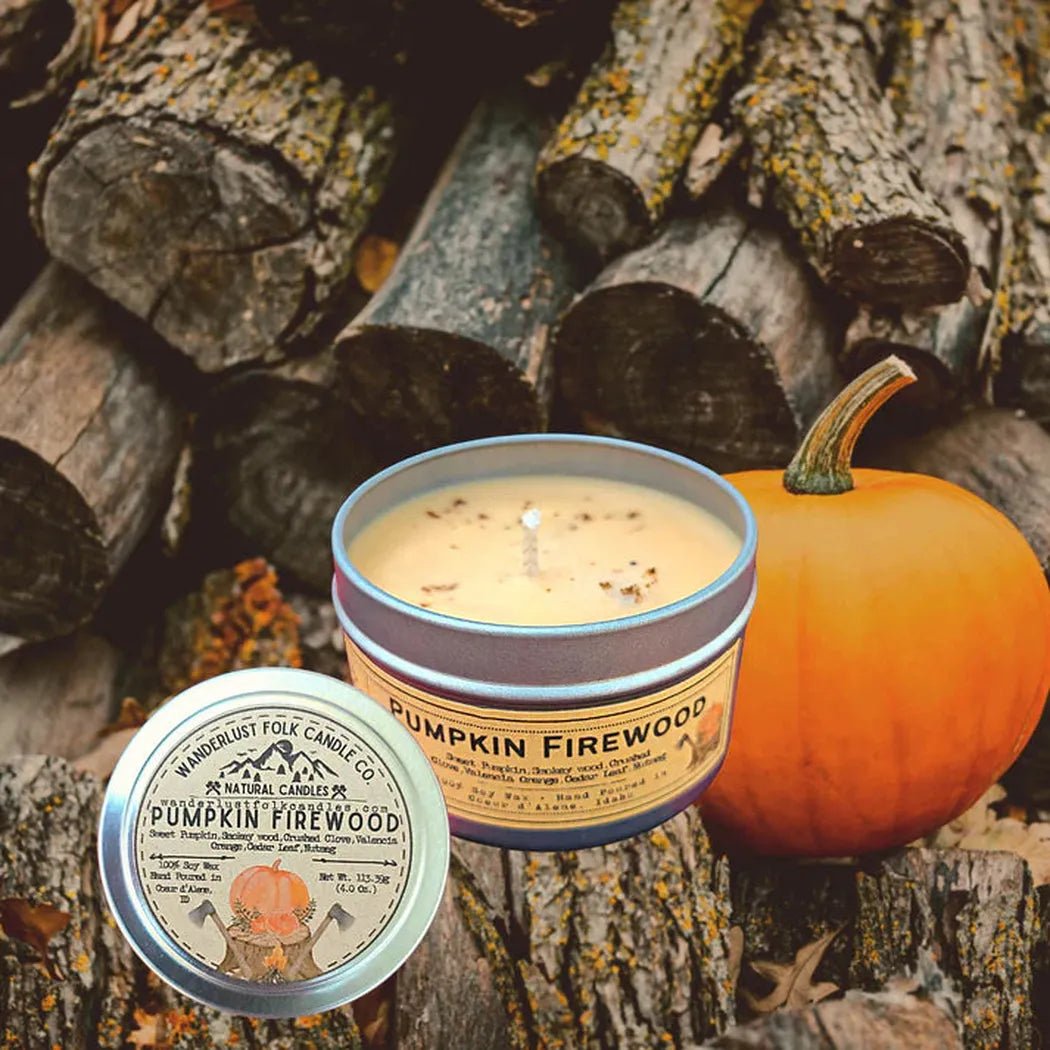 A close up of a Wanderlust Folk Pumpkin Firewood 4 oz candle tin and a pumpkin against a background of stacked firewood.