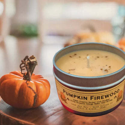 A 4 oz Wanderlust Folk Pumpkin Firewood candle tin sits on a wooden surface next to a small orange pumpkin.