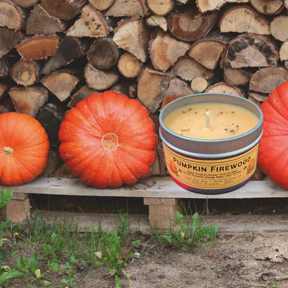 A 4 oz Wanderlust Folk Pumpkin Firewood candle sits on a wooden pallet with pumpkins and a woodpile background.