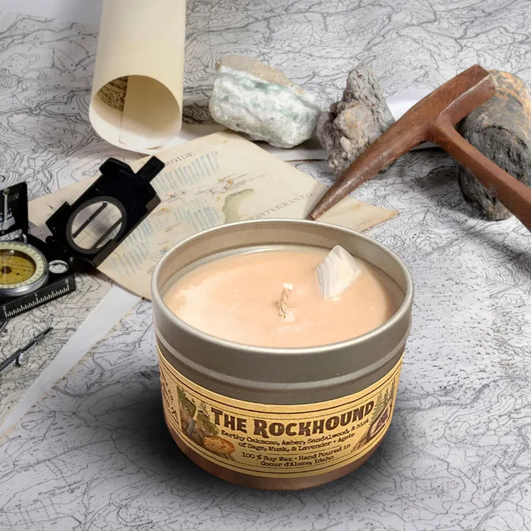 A 4 oz Wanderlust Folk The Rockhound candle sits on a map with a compass and pickaxe in the background.