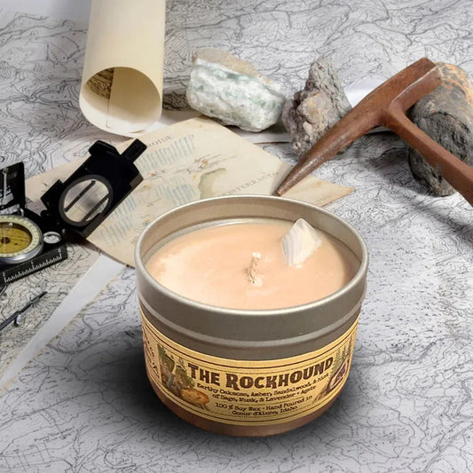 A 4 oz Wanderlust Folk The Rockhound candle sits on a map with a compass and pickaxe in the background.