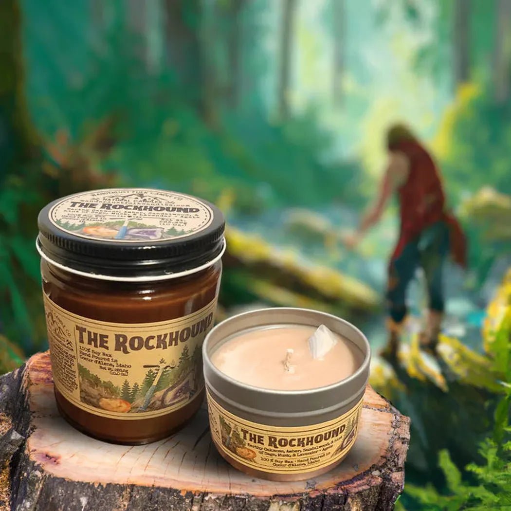 A 4 oz Wanderlust Folk The Rockhound candle and jar sit on a tree stump with a forest background.