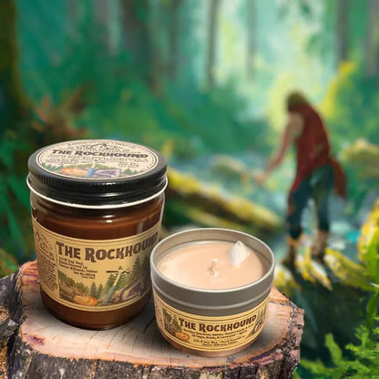 A 4 oz Wanderlust Folk The Rockhound candle and jar sit on a tree stump with a forest background.