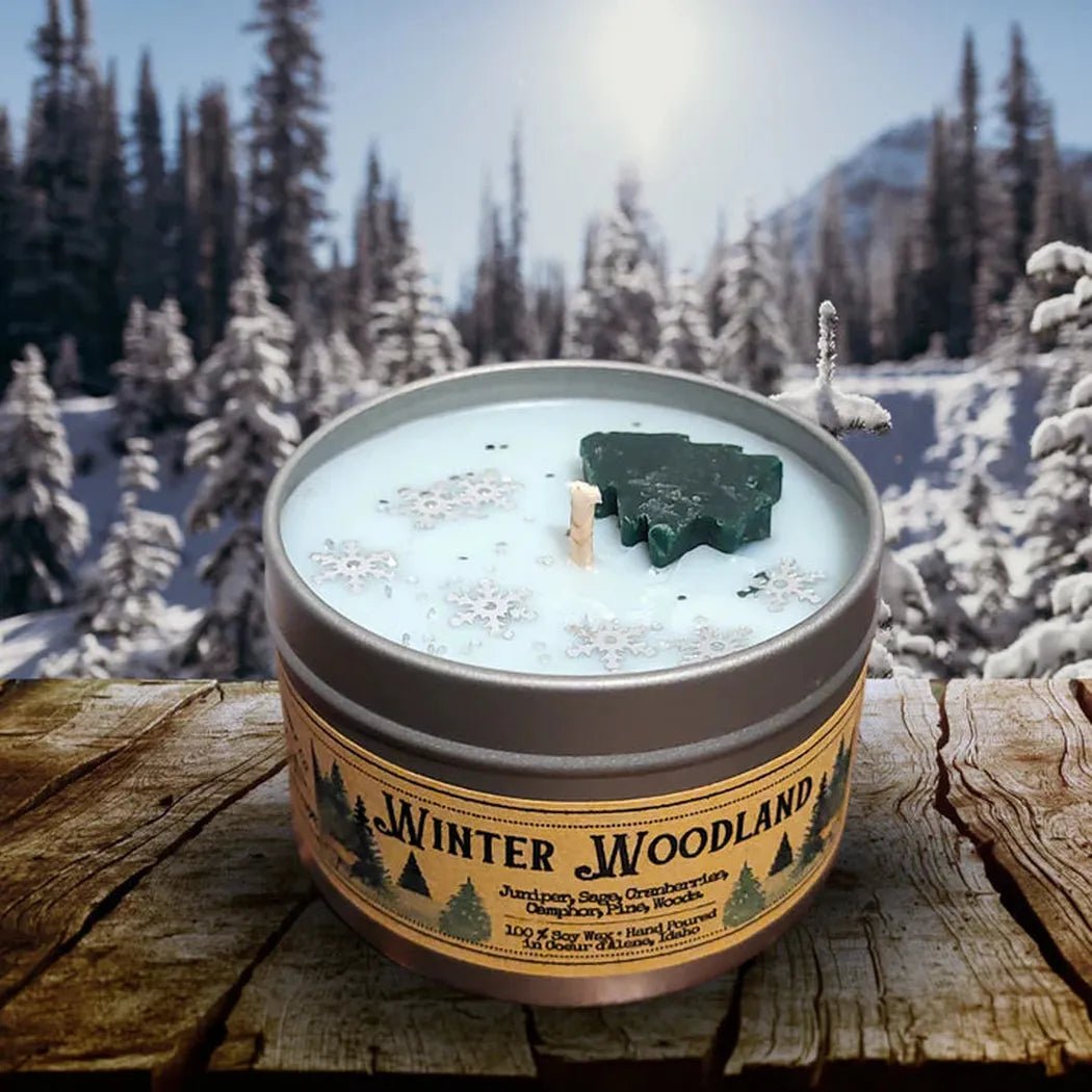 A 4 oz Wanderlust Folk Winter Wonderland candle in a tin sits on a wooden surface with a snowy forest background.