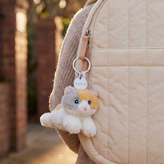 A close-up of the Warmies Calico Cat Keyring attached to a beige backpack strap, with a brick wall and outdoor path in the background.
