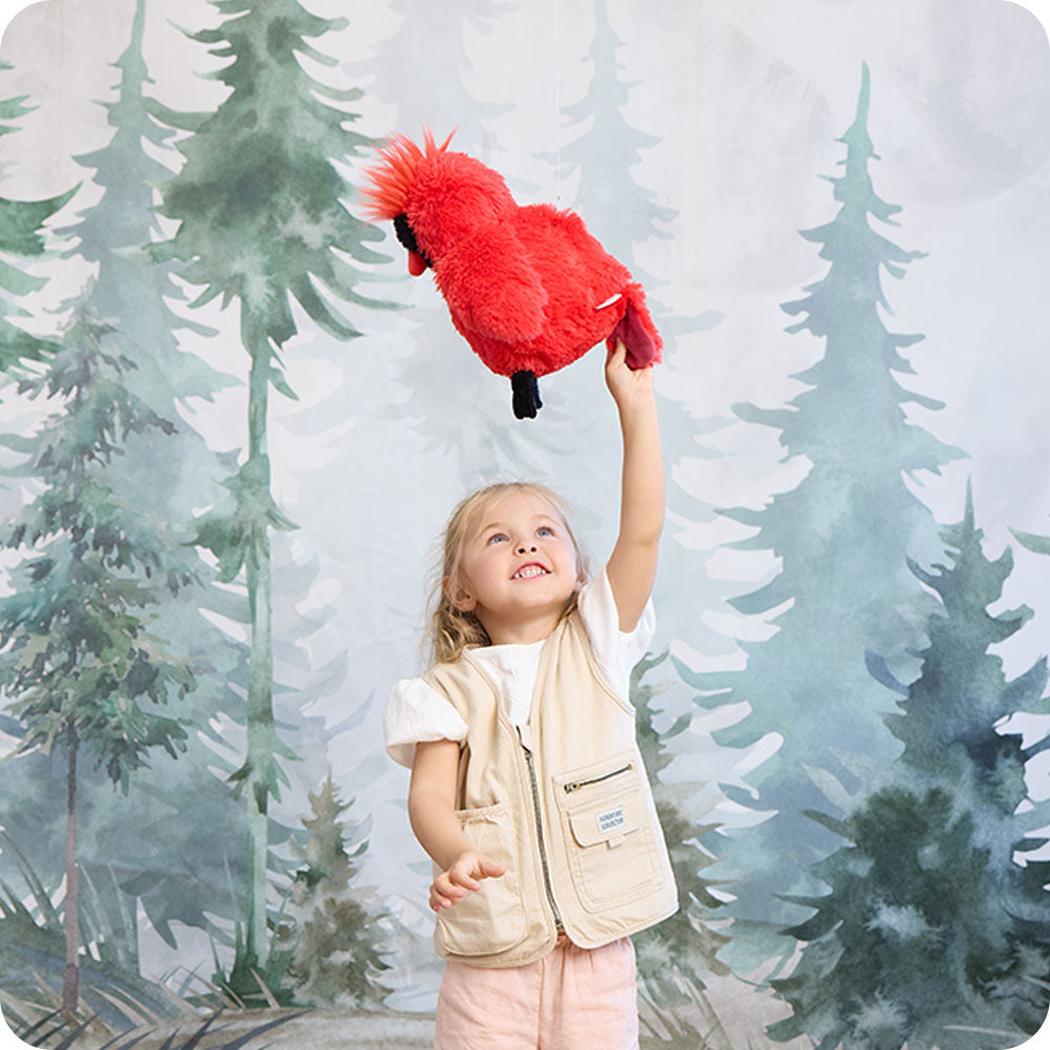 A young girl holds up the Warmies Cardinal plush toy against a forest backdrop.