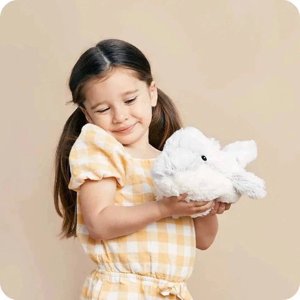 A young girl in a yellow and white checkered dress holds a white Warmies Dolphin plush toy against a beige background.