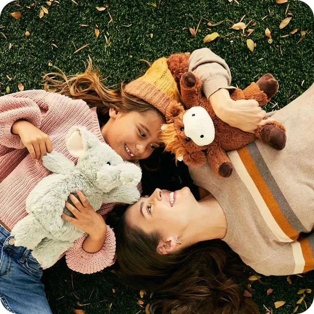 Two people lie on grass with a Warmies Highland Cow and a light blue plush toy.