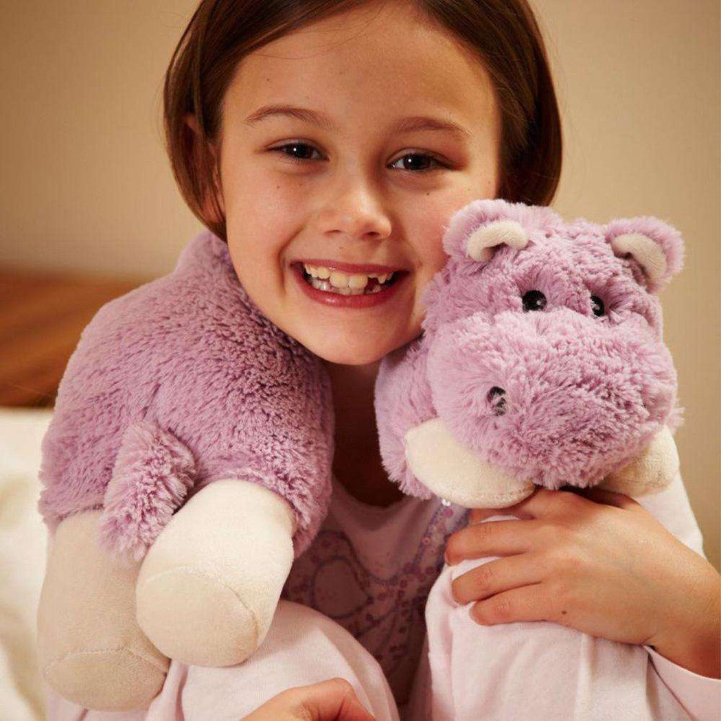 A young girl wearing a Warmies Hippo Cozy Wrap holds a matching purple stuffed hippo toy.