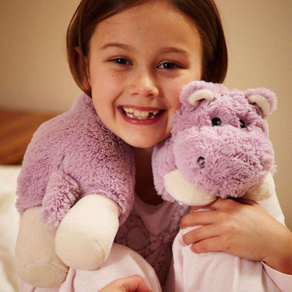 A young girl wearing a Warmies Hippo Cozy Wrap holds a matching purple stuffed hippo toy.