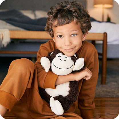 A young boy in brown pajamas sits on the floor holding a Warmies Monkey plush toy, with a bed and lamp in the background.