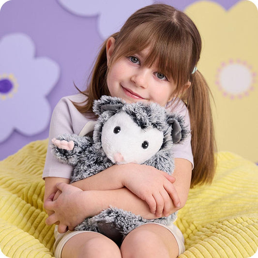 A young girl sits on a yellow blanket holding a Warmies Opossum plush toy against a purple and yellow floral background.