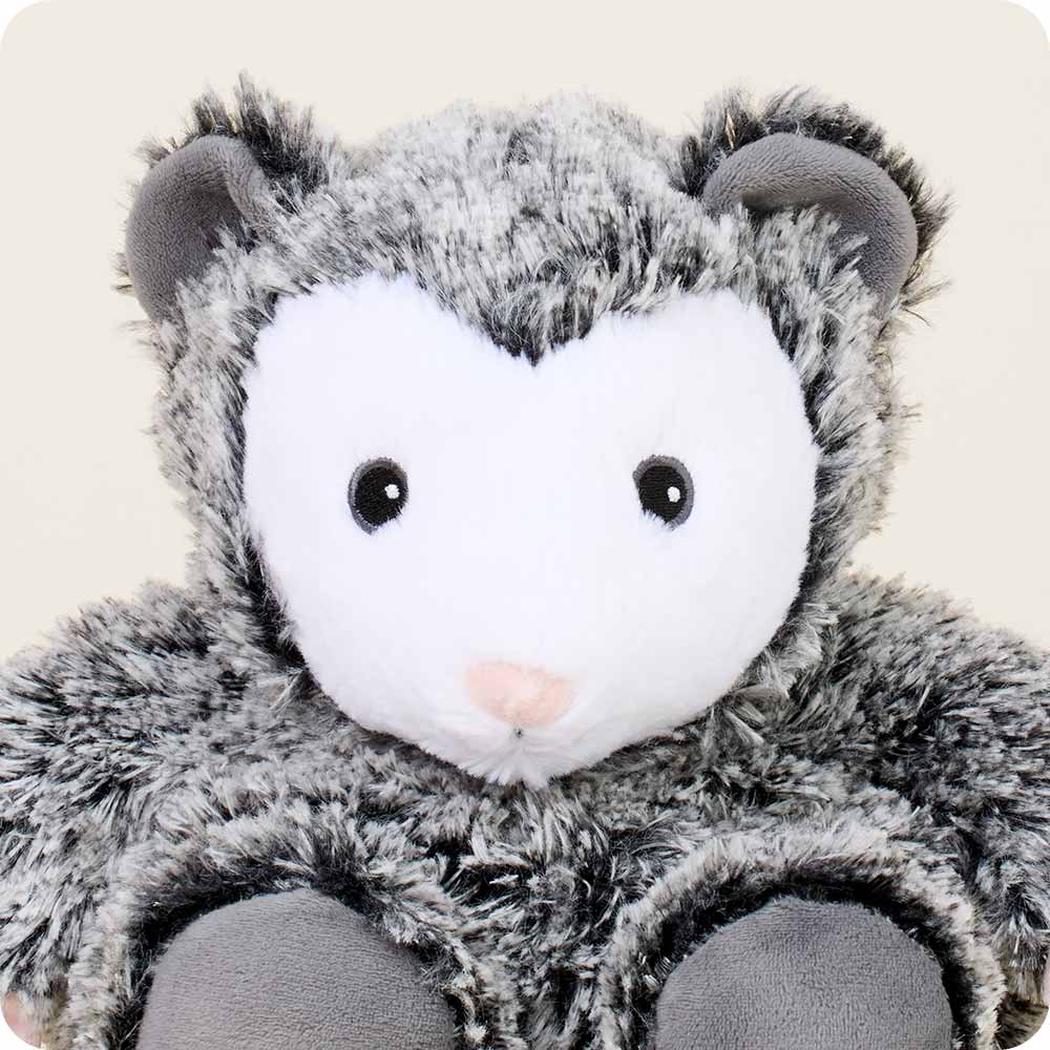 A close-up of the Warmies Opossum plush toy with a white face and charcoal fur against a light background.