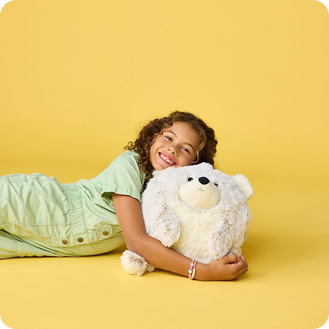 A girl in light green lies on a yellow background holding a Warmies Supersized Bear.