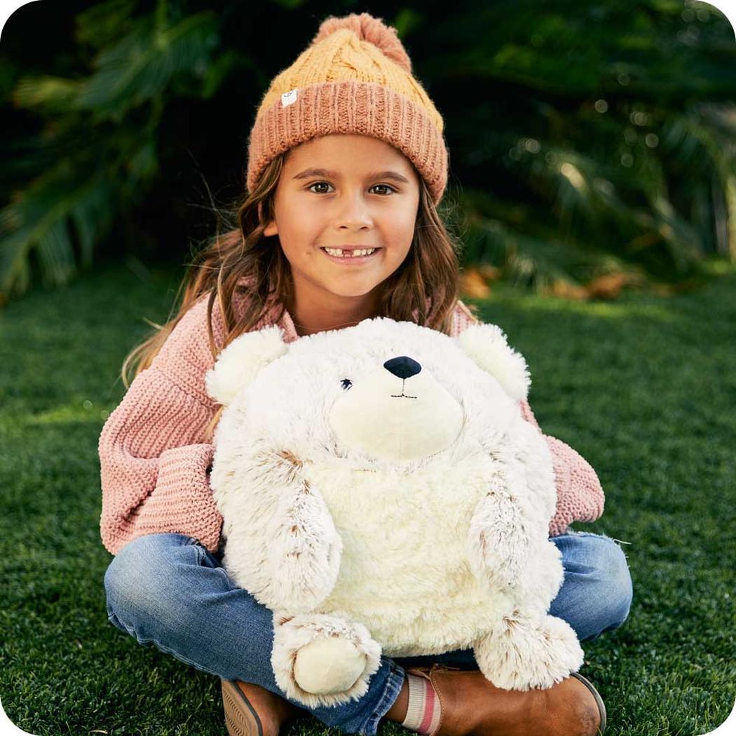 A girl sits on grass wearing a Warmies Supersized Bear plush toy.