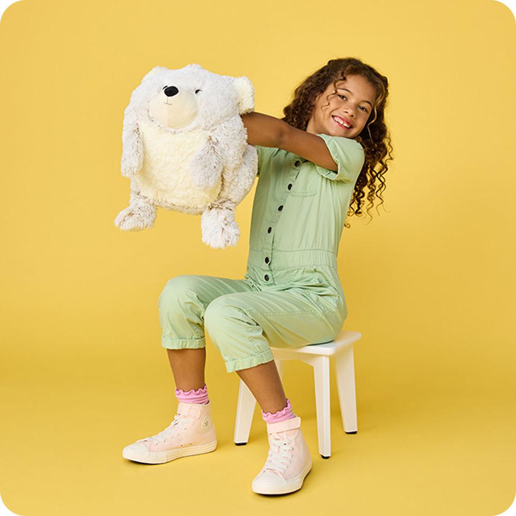 A girl in light green plays with a Warmies Supersized Bear on a white stool against a yellow background.