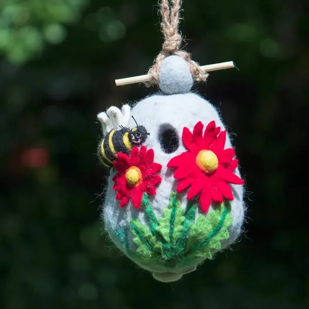 A hanging Wild Woolies Bumble Birdhouse with red flowers and a bee, set against a dark outdoor background.