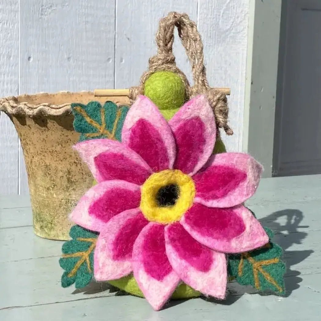A felt Wild Woolies Dahlia Star Birdhouse with pink petals and green leaves sits on a light blue surface near a weathered pot.