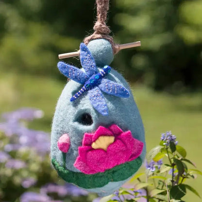A light blue Wild Woolies Lotus Dragonfly Birdhouse hangs from a rope with a blue felt dragonfly on top, set against a green outdoor background.