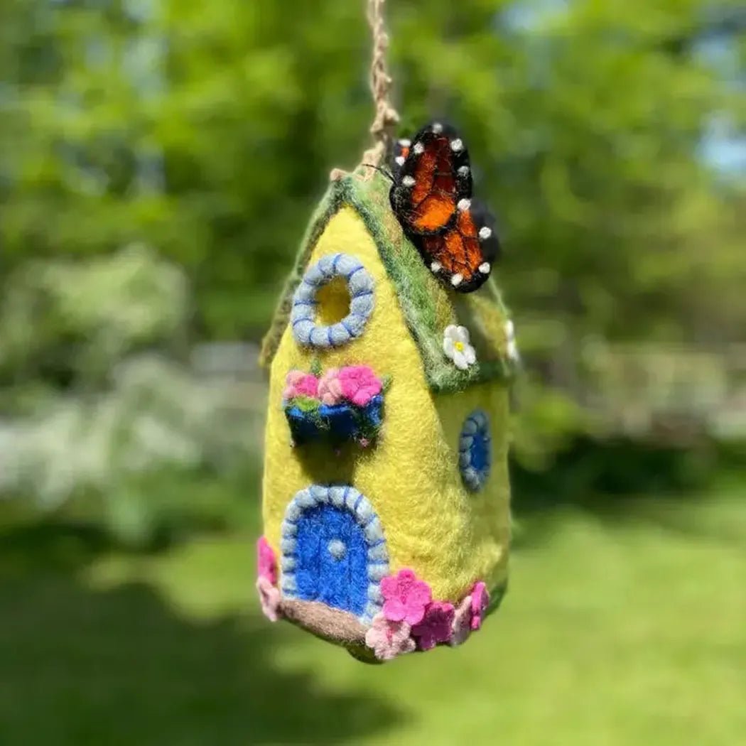 A hanging Wild Woolies Monarch Cottage Birdhouse with a monarch butterfly on top, set against a green outdoor background.