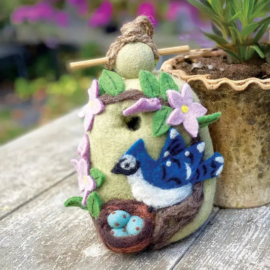 A close-up of the Wild Woolies Nesing Bluejay Birdhouse on a wooden surface with a potted plant in the background.