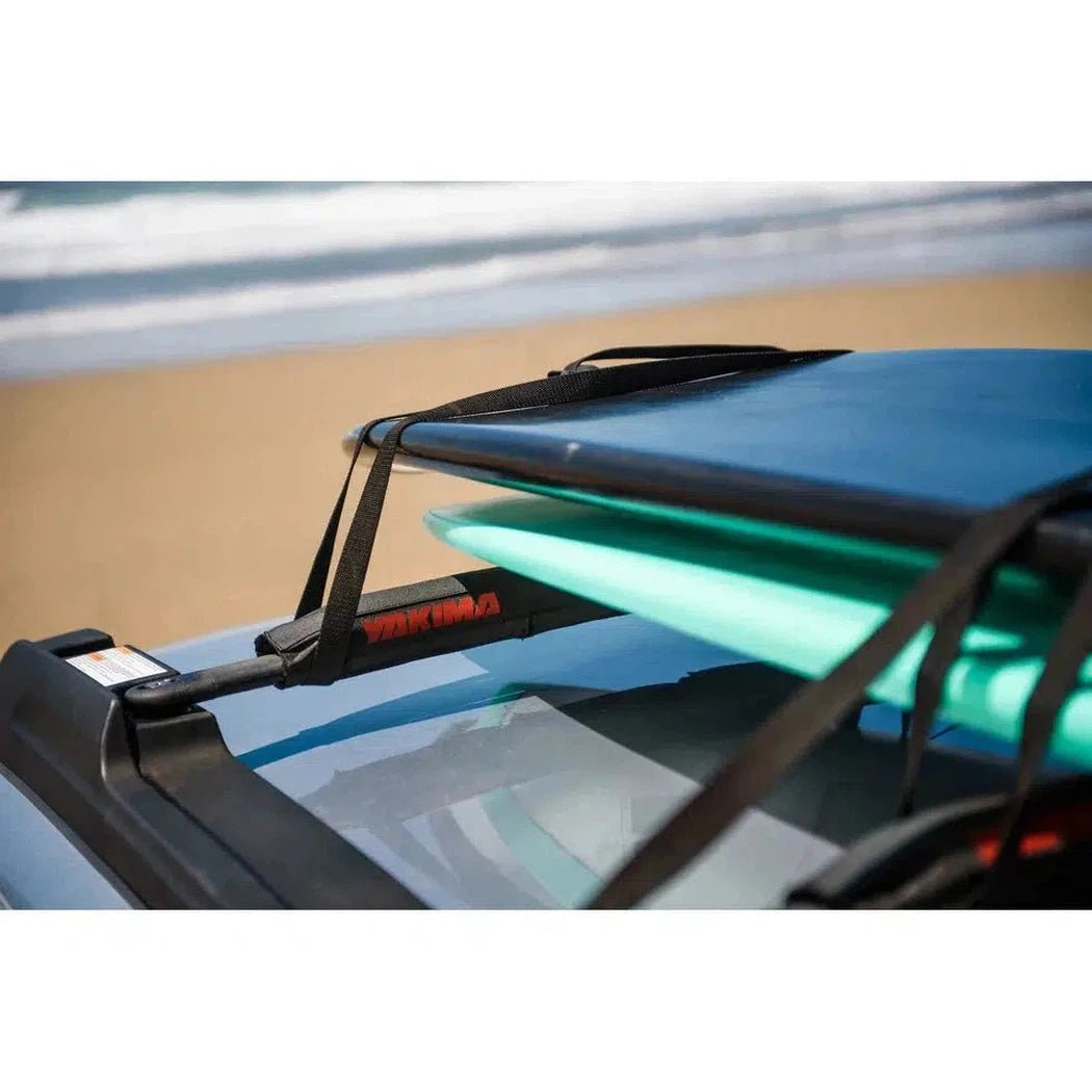 A close up of the Yakima Aero Crossbar Pads on a car roof rack with surfboards, showing the black straps and red label against a beach background.