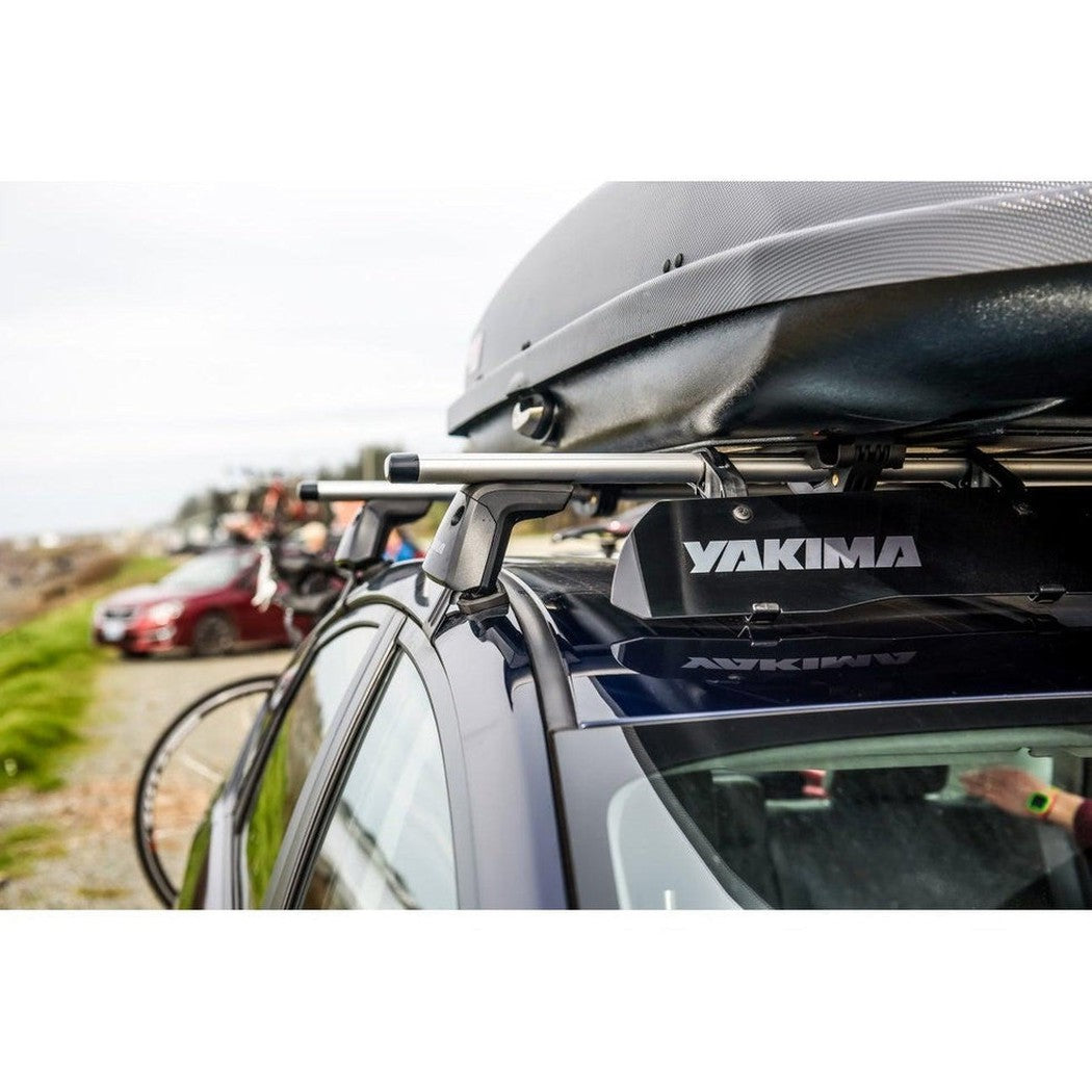 A close-up of a dark blue car with a Yakima roof rack and a black cargo box, showing a bicycle mounted on the side and a red car in the background.