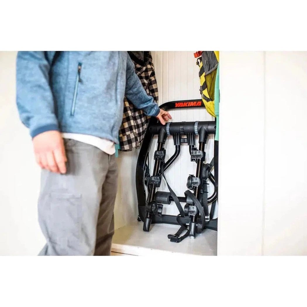A person in a blue jacket stands next to the Yakima FullBack bike rack in a white room with a checkered coat hanging nearby.