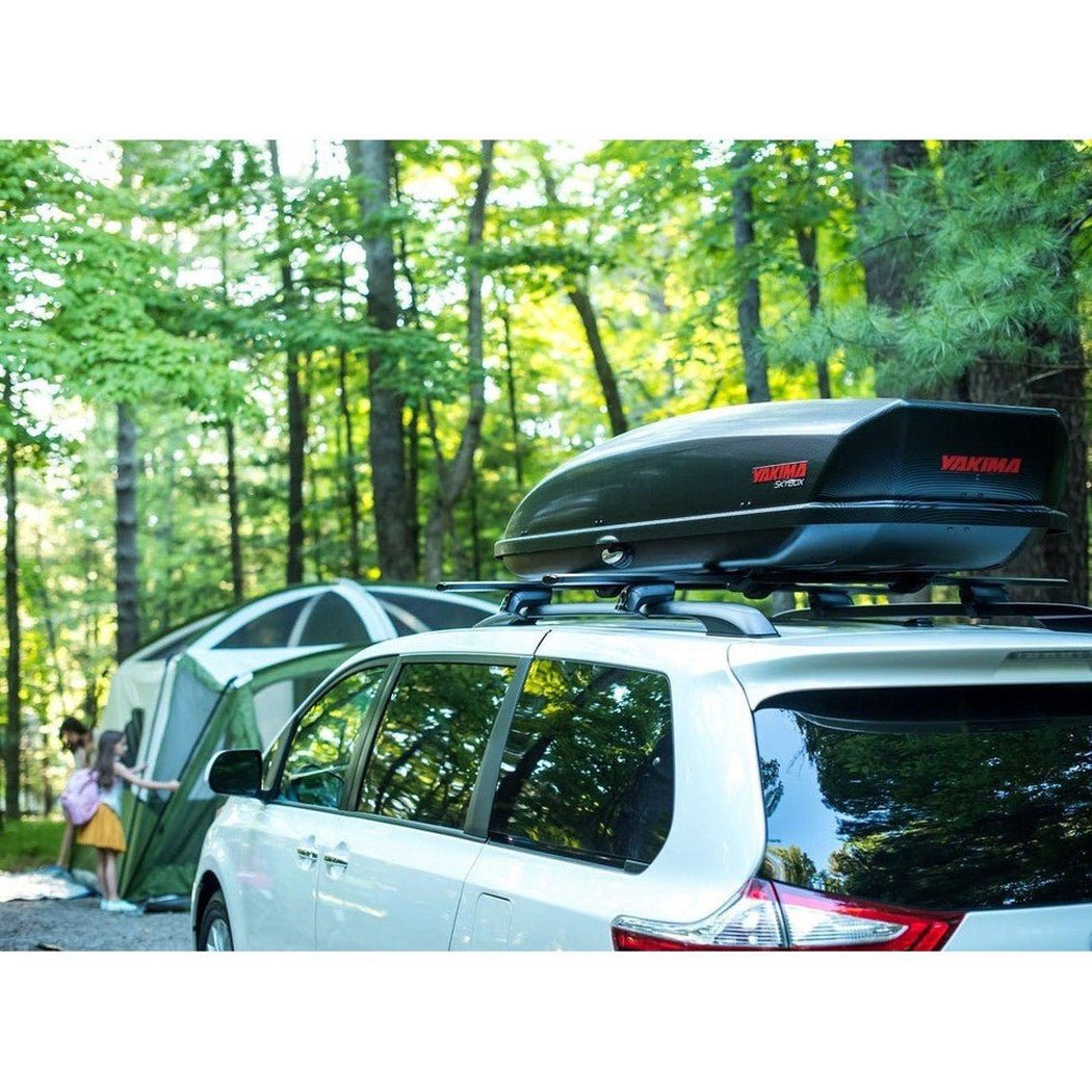 A white SUV with a black Yakima SkyBox 21 Carbonite cargo box on its roof sits in a wooded campsite with a tent and trees in the background.