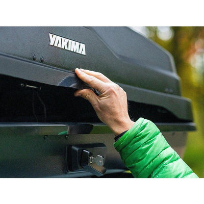 A hand opens the Yakima SkyBox NX 18 cargo carrier, showing its lock and the outdoor background.